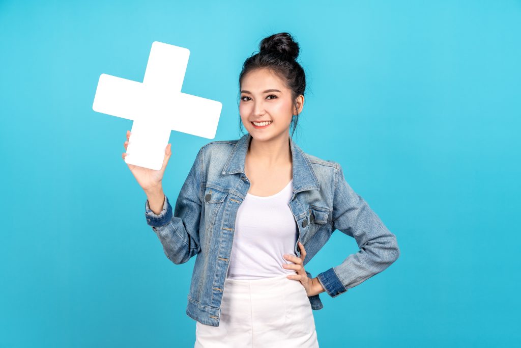 Happy asian woman smiling hand on hip and showing plus or add sign on blue background. Cute asia girl smiling wearing casual jeans shirt and showing join sign for increse and more benefit concept