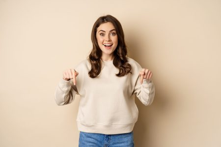 Enthusiastic attractive girl pointing fingers down, showing advertisement, link or promo below, standing over beige background.